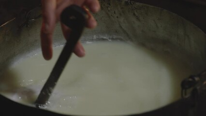 Stirring raw cheese curd in old iron cauldron with large metal ladle.