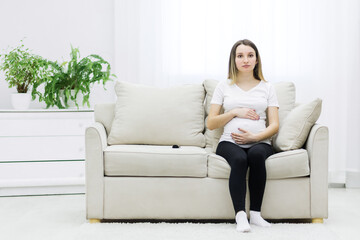 Pregnant woman sitting on white sofa and touching her stomach.