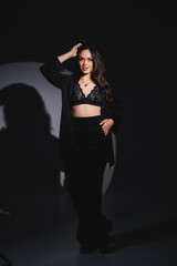 Fototapeta na wymiar A young beautiful woman in a black suit stands with her hands behind her head on a gray background. Attractive woman in underwear and a jacket