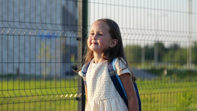Happy girl goes to school in preparatory form. Excited preschooler girl with backpack walks near sports-ground to study for first time slow motion, side view
