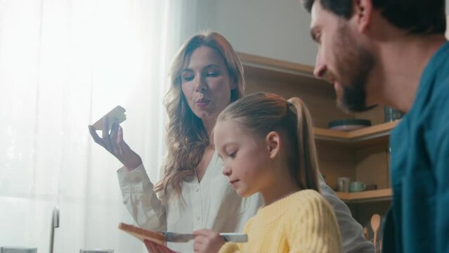 Moving shot Caucasian little girl child kid daughter spread chocolate paste on bread cooking breakfast for parents mother and father. Family eating lunch together at kitchen dining at table at home
