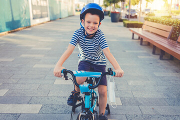 A smiling boy rides a bicycle on a city street and wears a protective mask attached to the bicycle. Healthcare concept. Portrait of confident boy sitting on bicycle