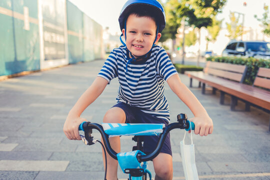 Cute Little Boy Child With Protective Medical Face Mask On The Handle And Safety Helmet Riding Bike In Sunny Summer Day At The Park During Covid-19 Outbreak, New Normal Lifestyle Concept.