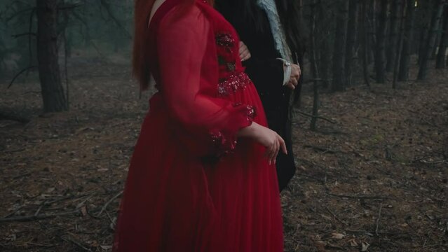 Goth man vampire in black tailcoat walking with gothic woman holding hand. fantasy couple two person back rear view. Red dress. Fog forest tree summer green nature. Guy with long dark hair. video 4k