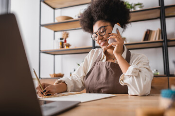 smiling african american craftswoman talking on smartphone and writing order in notebook near blurred laptop in workshop.