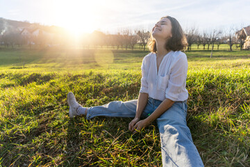 Young caucasian pretty woman in a white shirt sitting on the green grass enjoying the spring sun in...