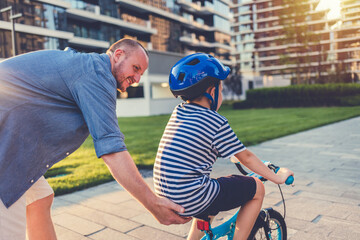 Little boy with cycling helmet getting support from his father when sitting on bike first time....
