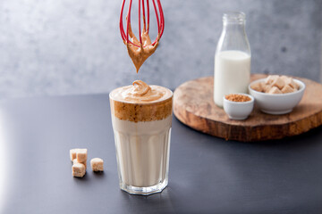 Dalgona korean coffee and a whisk with coffee foam with ingredients in the back: brown sugar cubes,...
