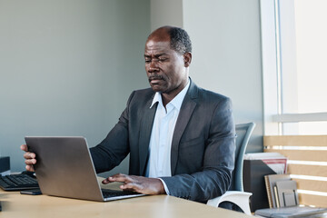 Experienced director of business company sitting by workplace in front of laptop in office and looking through online documents