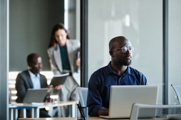 Pensive businessman in eyeglasses and blue shirt sitting by workplace in front of laptop and thinking of next point of business project