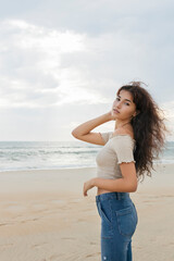 Young woman brunette teenager gir in jeans and top touching her long black hair posing and looking at camera in the beach on a cloudy day