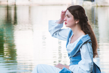 a woman with long hair in a vintage historical blue dress sits by the river and looks at the...