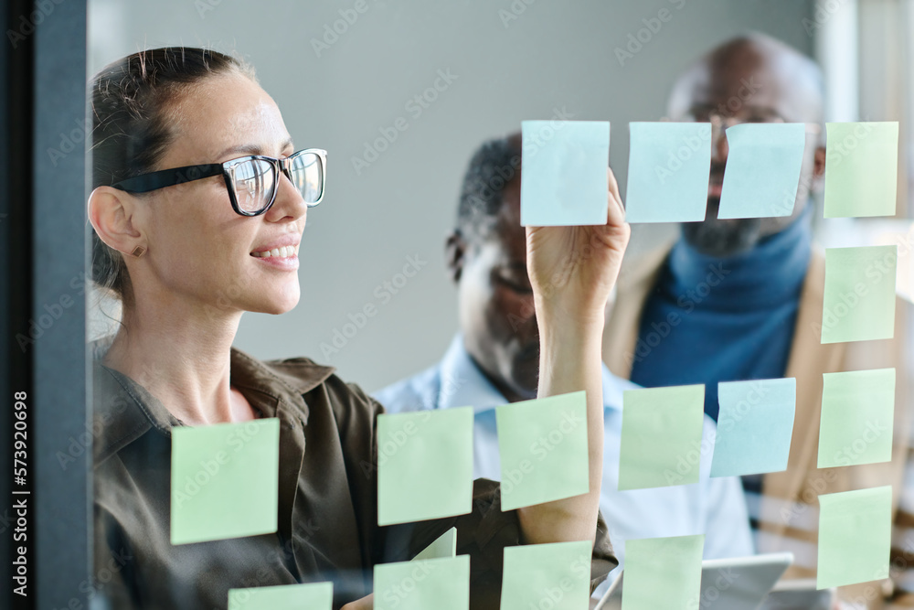 Poster young business coach in eyeglasses making notes on notepaper while preparing for presentation of com