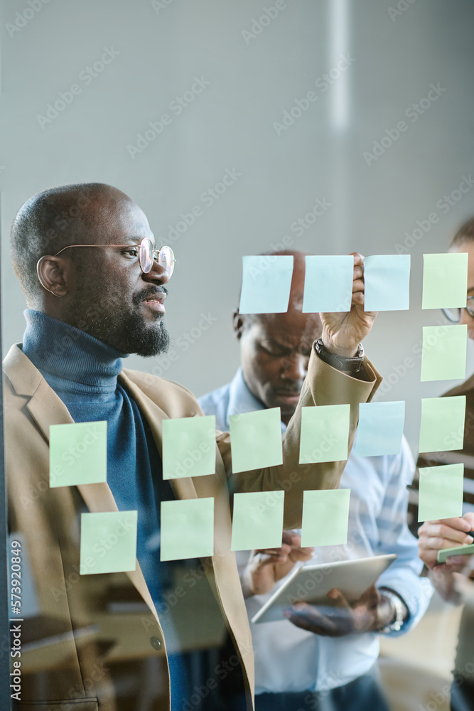 Poster young confident businessman in casualwear writing down tasks on notepapers while standing in front o