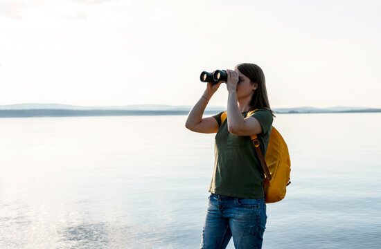 Young woman with yellow backpack looking through binoculars at birds on lake or river Birdwatching, zoology, ecology Research in nature, observation of animals Ornithology wildlife manager copy space