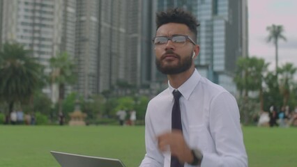 Young businessman in wireless earphones sitting on green lawn in city park, typing on laptop and using smartphone while working remotely outdoors on summer day - Powered by Adobe
