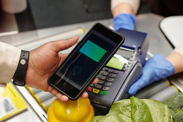 Close-up of consumer hand holding smartphone over card reader during contactless payment for food products in supermarket