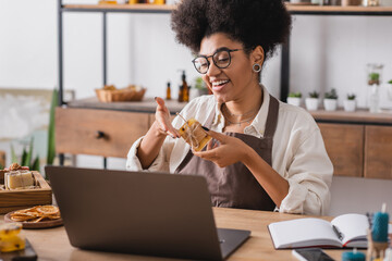 happy african american woman in eyeglasses showing handmade soap during video call on laptop in craft workshop.