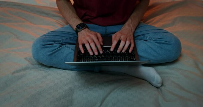 A man is typing on a laptop, standing on his feet, sitting in a lotus position on the bed, Close-up of male hands on the keyboard, no face, front view from above