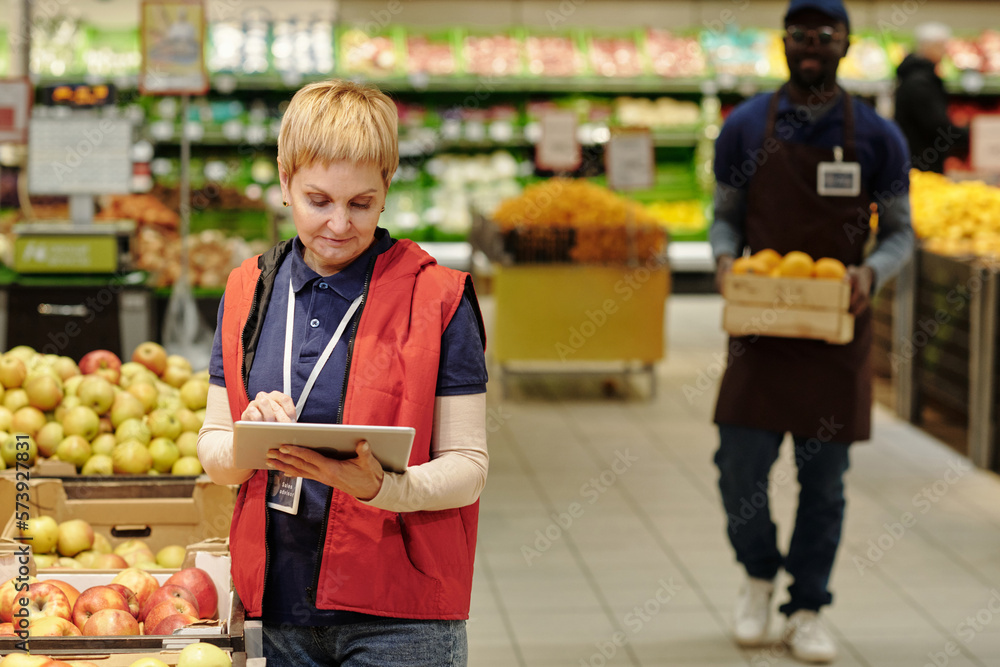 Poster mature blond female administrator of supermarket in uniform standing by large display with fresh app
