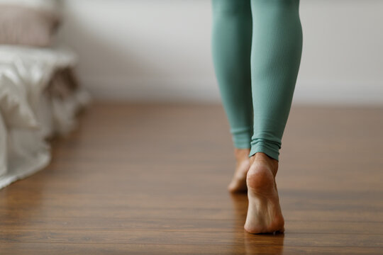 Young woman in sports leggings walks barefoot on the warm floor in the bedroom. Close-up of cropped image of barefoot girl. Underfloor heating system wood panels.