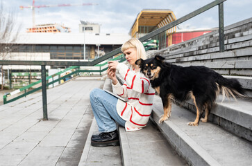 Young freelance expat woman sitting on the stairs of office building drinking coffee and working on laptop computer with her dog because it's bring-your-pets-to-work day. Web designer working outdoor