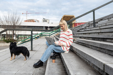 Young freelance expat woman sitting on the stairs of office building drinking coffee and working on laptop computer with her dog because it's bring-your-pets-to-work day. Web designer working outdoor