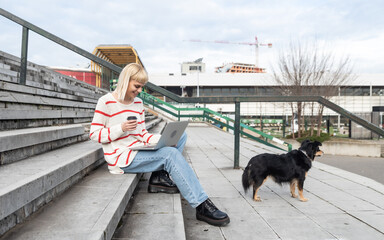 Young freelance expat woman sitting on the stairs of office building drinking coffee and working on laptop computer with her dog because it's bring-your-pets-to-work day. Web designer working outdoor