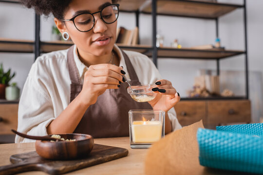 african american craftswoman in eyeglasses heating beeswax above burning candle near wooden bowl and chopping board.