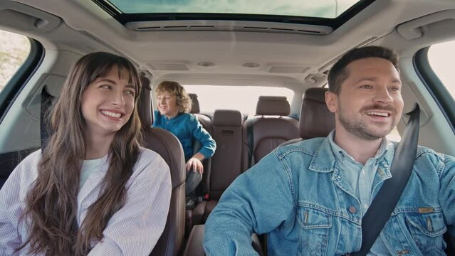 Portrait of lovely happy young family inside car. Handsome cheerful dad driving car with beautiful loving mom and cute little blonde son having fun sitting in baby car seat. Car trip concept.