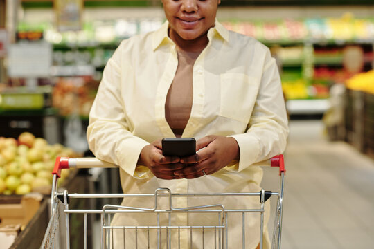 Close-up Of Young Black Woman Scrolling In Smartphone While Walking Along Grocery Department And Pushing Shopping Cart