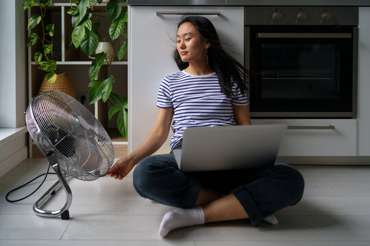 Work From Home In Heatwave. Happy Smiling Asian Girl Remote Worker Working Online On Laptop, Sitting On Floor Near Electric Fan At Home, Enjoying Fresh Air, Keeping Home Office Cool In Summer Heat