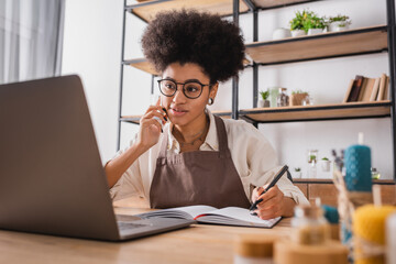 positive african american artisan with notebook and pen accepting order on smartphone near laptop.