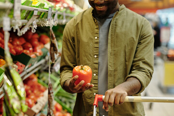 Close-up of happy young male consumer holding red ripe fresh pepper while standing by display with vegetables in supermarket