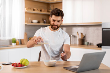 Fototapeta na wymiar Smiling adult caucasian male in white t-shirt pours milk into cereal at table with laptop, watching video
