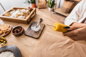 partial view of african american craftswoman holding handmade candle near parchment and natural ingredients on wooden table.