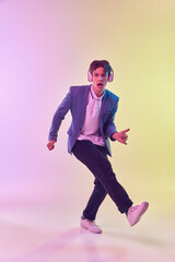 Full-length portrait of young man in headphones dancing isolated on gradient yellow-purple background in neon light. Concept of human emotions, youth culture.