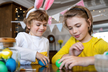 Easter Family traditions. Two caucasian happy children with bunny ears dye and decorate eggs with paints for holidays playing together. Kids having fun.