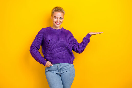 Photo of charming smiling blonde short hair girl wear violet knitted jumper hold palm promo new cheap product isolated on yellow color background