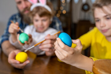 Fototapeta na wymiar Easter Family traditions. Father and two caucasian happy children with bunny ears dye and decorate eggs with paints for holidays having fun together at home. Negative space