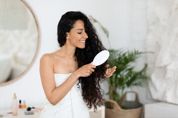 Happy millennial caucasian brunette lady in towel combing long curly hair, enjoy spa treatment
