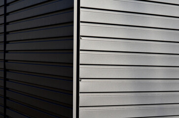 gray building is lined with square panels. metal sheet wall cladding with scalloped design. corrugated sheet, dark gray, riveted. horizontal stripes, shadows, corner, fence, aluminium, sheet