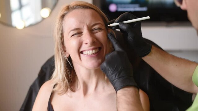 Man's hands in black gloves hold pencil and show on woman's face places for cosmetic injections. Make botox and hyaluronic acid in  beauty salon against wrinkles near of eyes.
