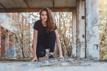 Curly-haired teenage girl in black jeans squats on abandoned construction. Vacation outside city.
