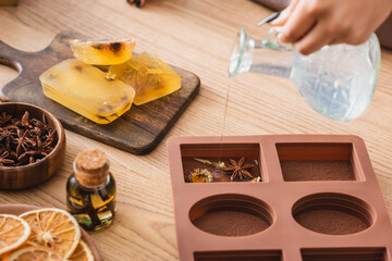 partial view of african american craftswoman pouring liquid soap in silicone mold with dried herbs near essential oil and orange slices.