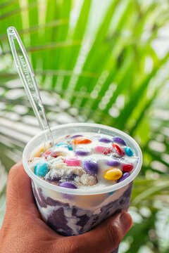  vertical photo of a hand holding an creamy acai bowl with condensed milk and colorful chocolate chip with background of palm trees