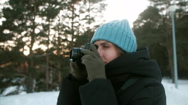 Attractive young woman photographer in blue hat, eyeglasses and black jacket, photographing on digital photo camera at winter time in the park covered snow. Slowmotion.