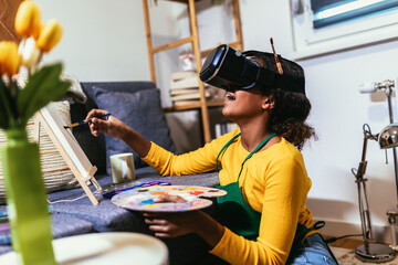 Happy black school girl painting with a paintbrush at home using VR glasses to immerse herself in...