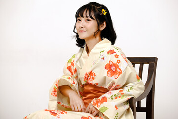 Fototapeta na wymiar Beautiful young woman smiling and wearing traditional Japanese casual summer kimono or yukata, looking at camera on a chair with white background