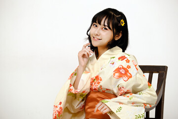 Beautiful young woman smiling and wearing traditional Japanese casual summer kimono or yukata, looking at camera on a chair with white background